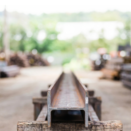 Close-up of the end of a steel H-beam resting on wooden blocks in an industrial storage yard, with the background heavily blurred.